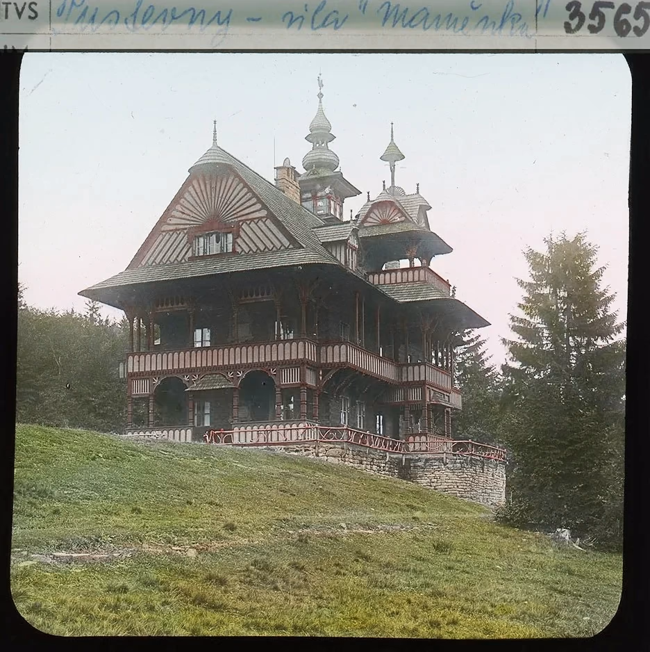 Kolorovaný skleněný diapozitiv kolem roku 1910: útulna Maměnka na svahu, dřevěná stavba s pavlačemi a vížkami, v popředí louka; na destičce dobový popisek „Pustevny – vila Maměnka“