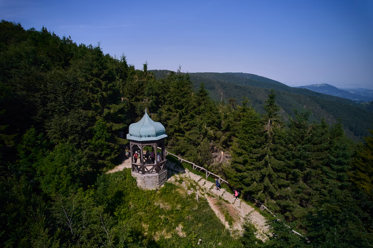 Letecký pohled na altán s cibulovou střechou na zalesněném hřebeni, k němu vede ohrazená pěšina, po které jdou turisté