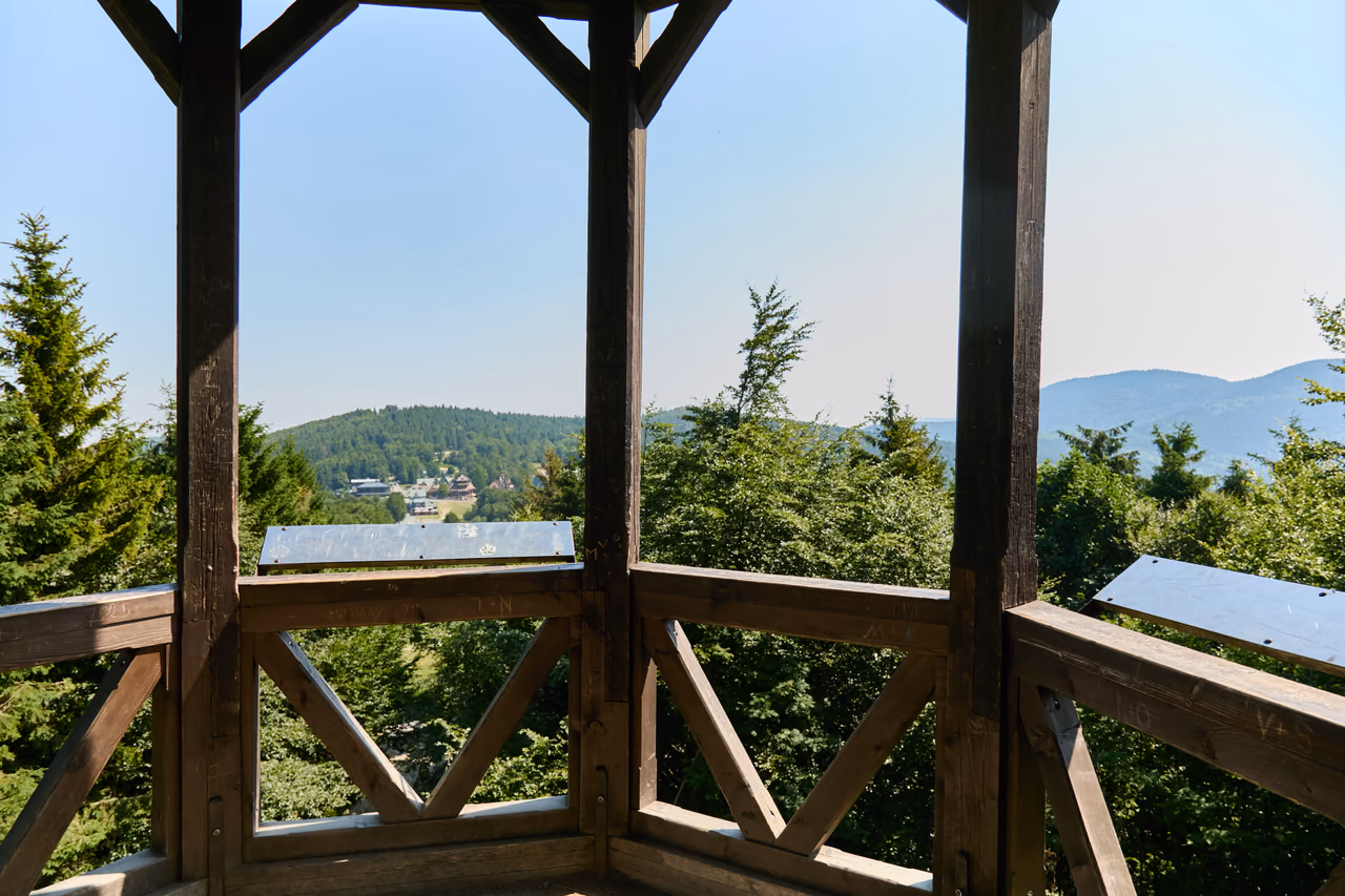 Pohled zevnitř altánu mezi dřevěnými trámy přes zábradlí na zalesněné kopce a osadu v údolí, na zábradlí dvě kovové desky s panoramatickou mapou