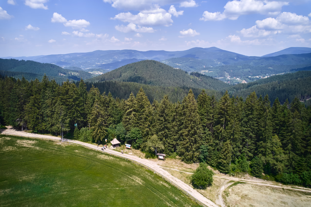 An aerial view of a ridge meadow with a wooden shelter at the edge of the spruce forest, the wooded ridges of the Beskids behind