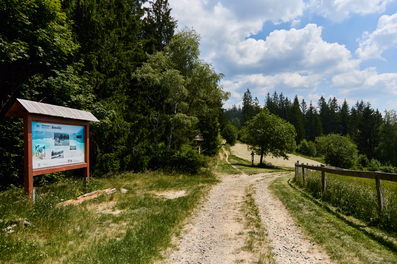 A wooden information panel reading Benešky by a gravel track, a fenced pasture and meadow on the right, wood behind