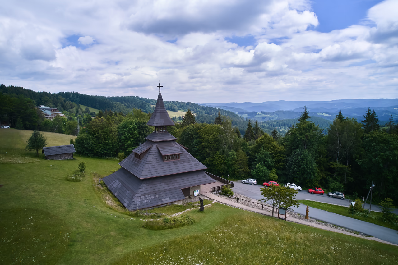 A close aerial view of the Soláň bell tower – a stepped shingle roof with a cross, visitors on the terrace by the entrance