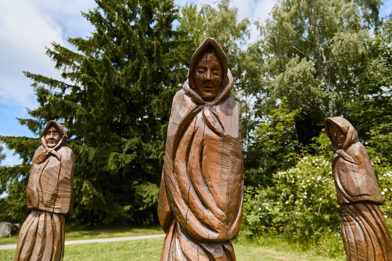 A wooden sculpture of a figure wrapped in a shawl, trees behind