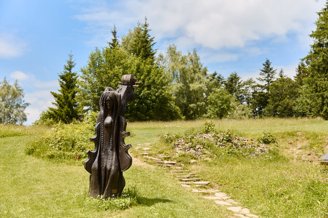 A dark wooden sculpture in the shape of a violin on a meadow, a stone path around it