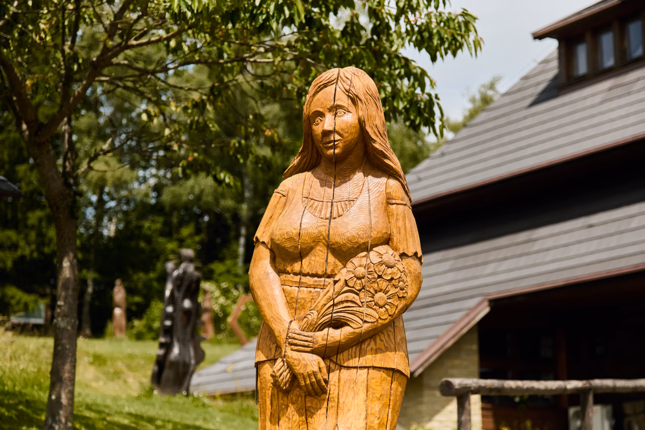 A pale wooden sculpture of a woman holding flowers by a shingled building