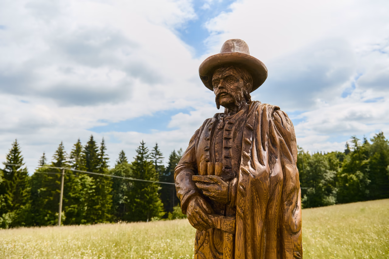 A wooden sculpture of a man in a hat and long cloak on a mountain meadow by Čarták