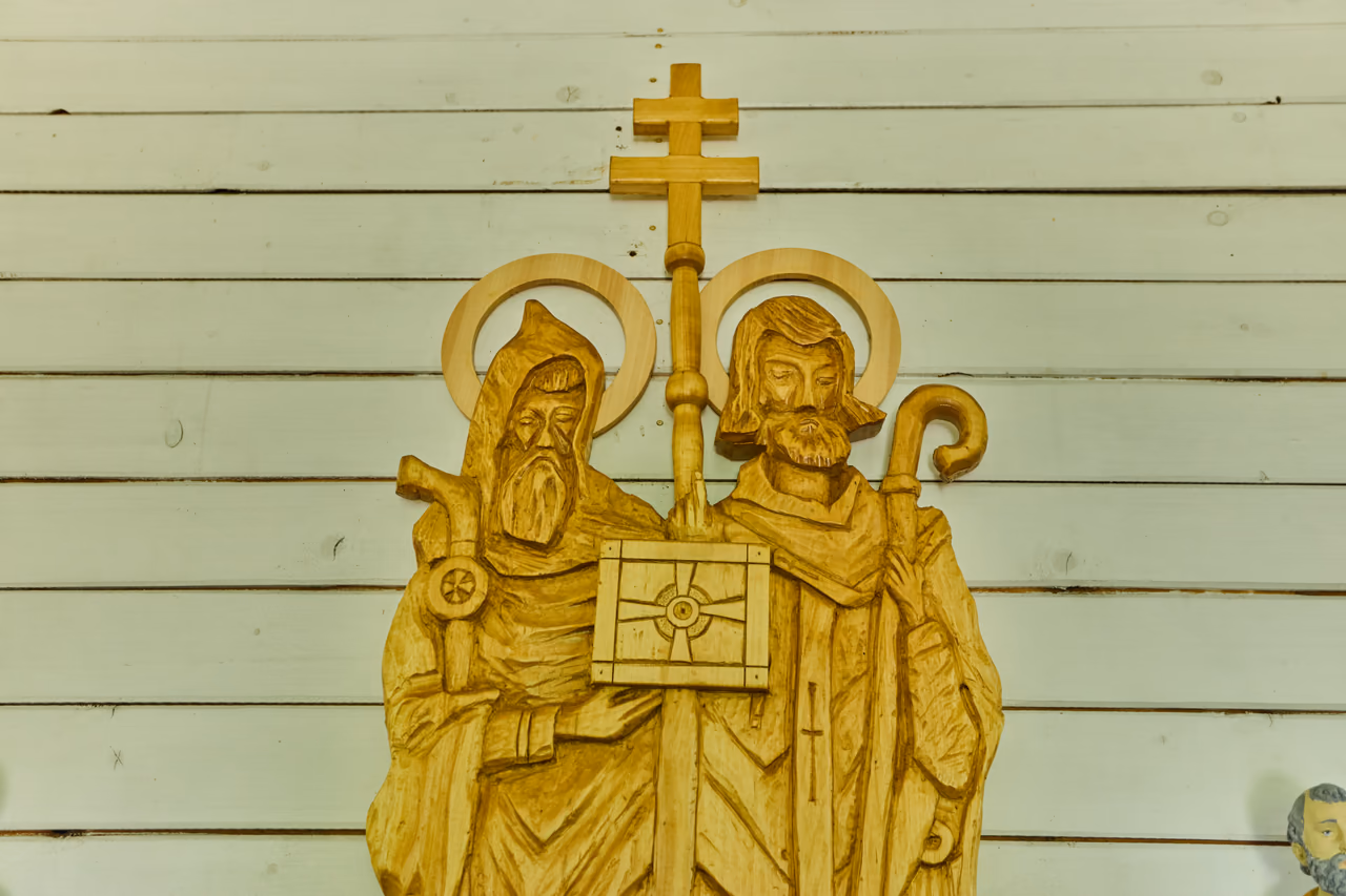 Detail zlaceného dřevěného reliéfu na bílé prkenné stěně: dvě postavy se svatozáří, vlevo mnich, vpravo biskup, oba se zdobenou berlou, mezi nimi dvojramenný kříž a kniha, kterou drží společně