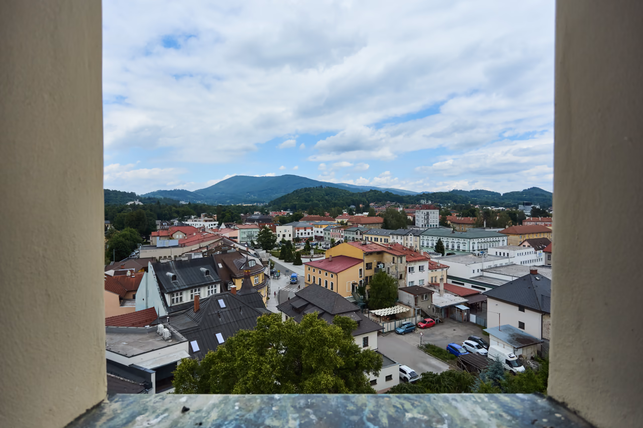 Výhled z okna kostelní věže na střechy centra Rožnova a zalesněné kopce v pozadí
