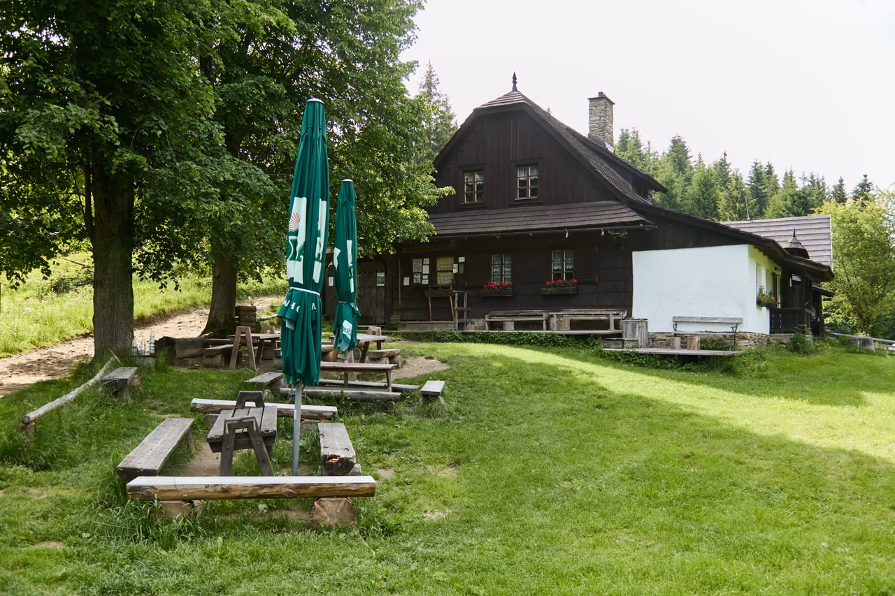 Pohled přes travnatou louku ke chalupě, v popředí dřevěné stoly s lavicemi a složené slunečníky