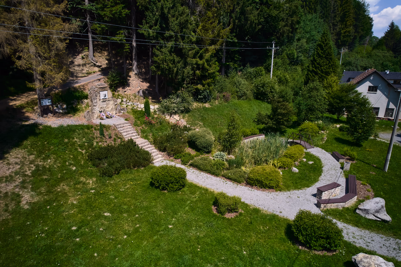 An aerial view of the memorial on the slope above the road: stone steps, a landscaped garden with paths and benches, a family house and the road on the right, spruce forest behind the memorial