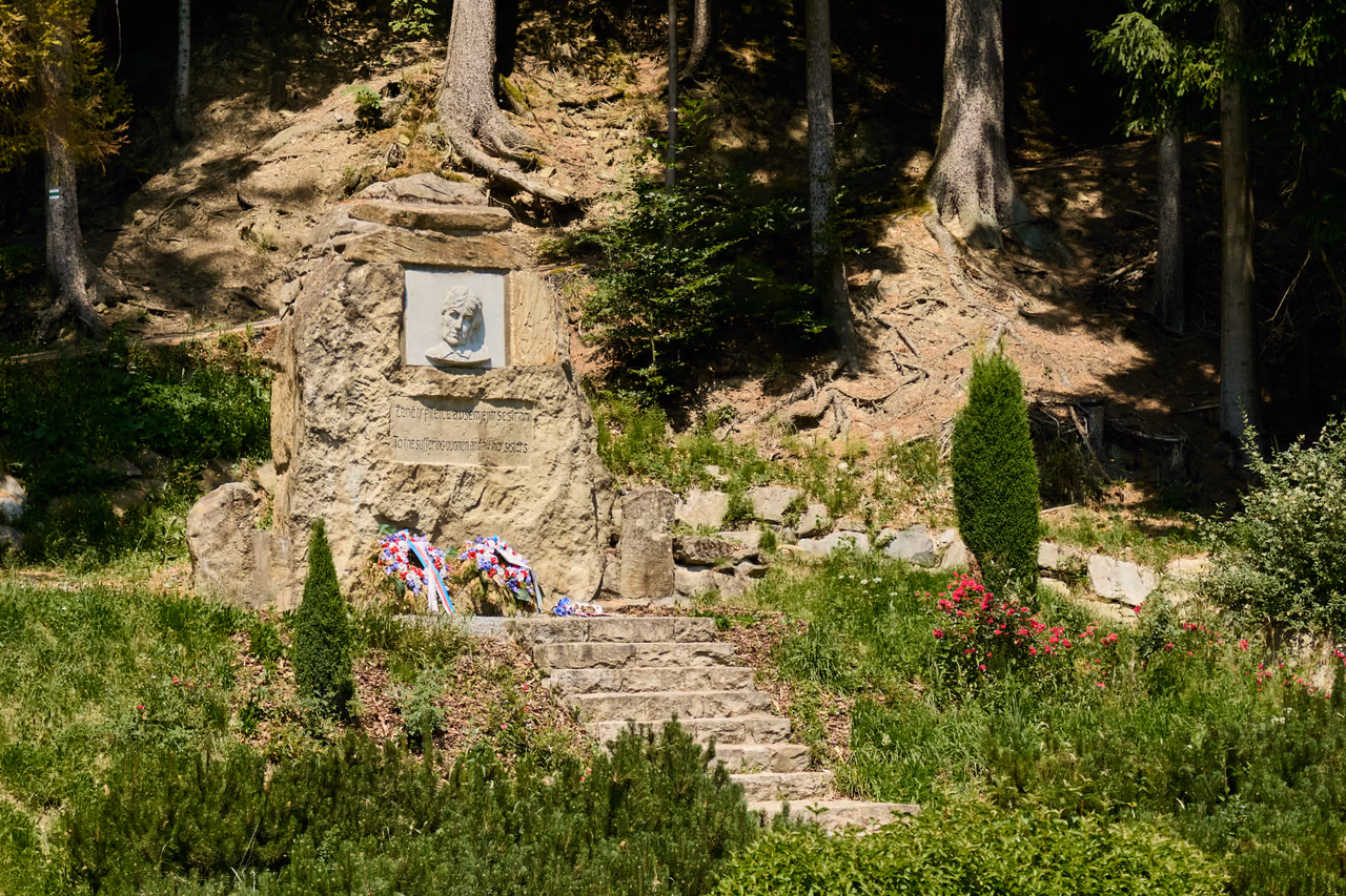 A sandstone boulder on the slope above stone steps, wreaths with ribbons in the national colours at its foot, low ornamental shrubs and flowering perennials around it