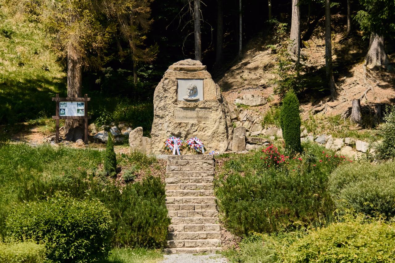 The memorial on the slope with an information panel on the left, stone steps in the middle, spruce forest behind the boulder