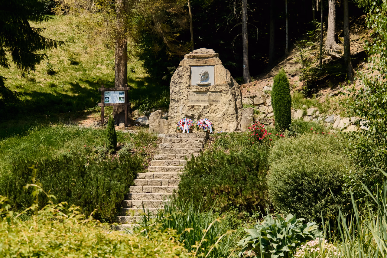 A view of the boulder from the right: stone steps between clipped shrubs, the memorial above them under the tree canopy