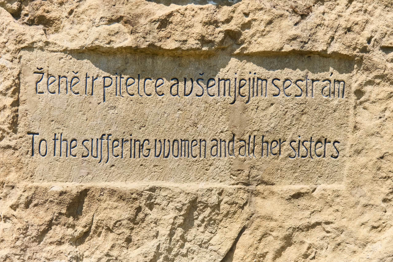 A close-up of the bilingual inscription carved into sandstone: “Ženě trpitelce a všem jejím sestrám” and the English rendering of the same sentence below it