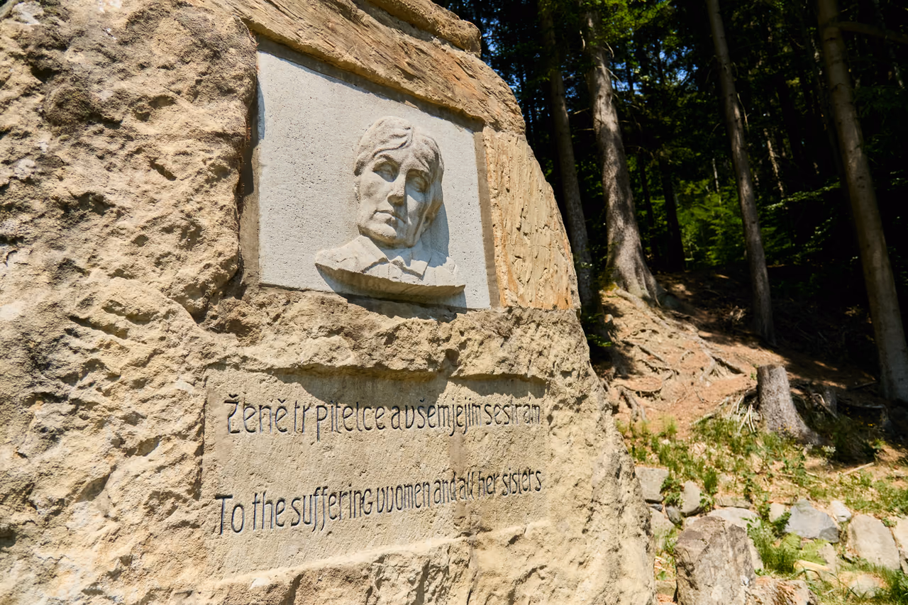 The face of the sandstone boulder with a plaque bearing a relief and a bilingual inscription carved below it, rough stone around it and spruce forest behind