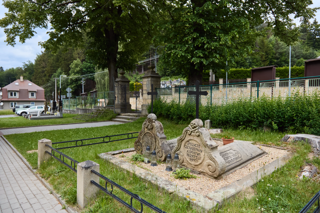 Pohled od chodníku: dvě tumby s křížovými náhrobky na štěrkovém záhonu v trávníku, za nimi vzrostlá lípa a kamenná brána hřbitova