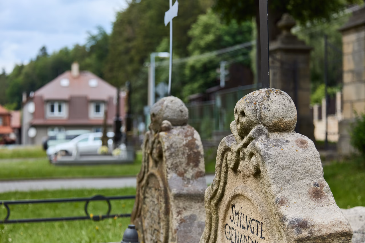 Zadní pohled na obě kamenné lebky s litinovými kříži, v rozostřeném pozadí domy obce a silnice