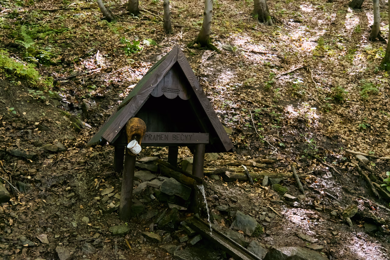 Hölzernes Schutzdach über der Quelle von vorn, mit der eingeschnitzten Aufschrift PRAMEN BEČVY und einem Becher, das Wasser fließt durch einen Holztrog den Hang hinab