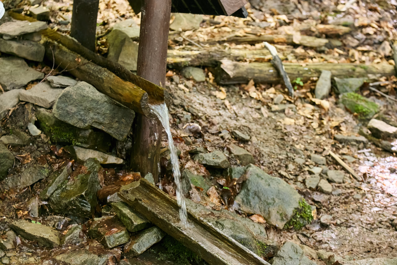 Detail des Endes der Holzrinne, aus der ein Rinnsal auf die Steine und das nasse Holz darunter fällt