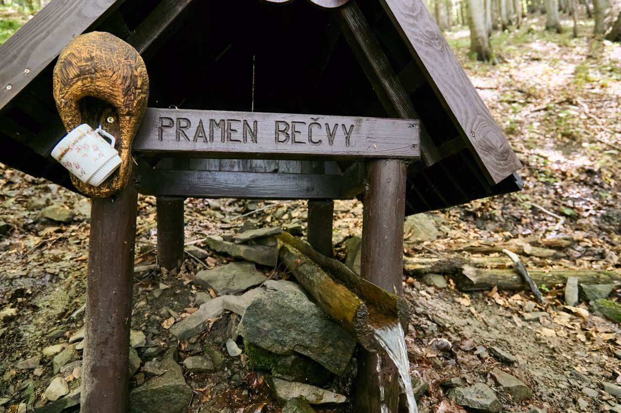 Stirnseite des hölzernen Schutzdachs mit der eingeschnitzten Aufschrift PRAMEN BEČVY, links ein geschnitzter Holzkopf, an dem ein Porzellanbecher hängt; unter dem Dach fließt Wasser aus der Rinne
