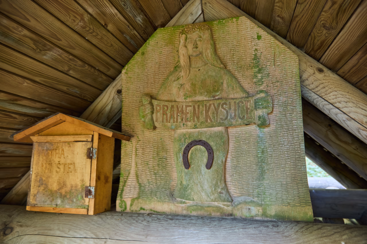Vyřezávaná dřevěná deska s ženskou postavou v koruně a nápisem Pramen Kysuce, pod nápisem přibitá podkova; vlevo malá dřevěná skříňka s dvířky