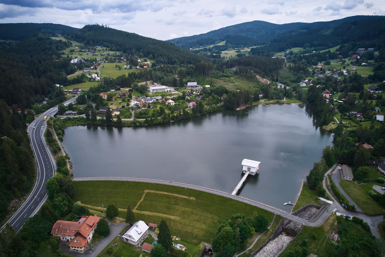 Luftaufnahme des Bogens des Erddamms mit dem Turmbauwerk und dem Steg, links die Straße I/35, rechts die Schussrinne der Hochwasserentlastung