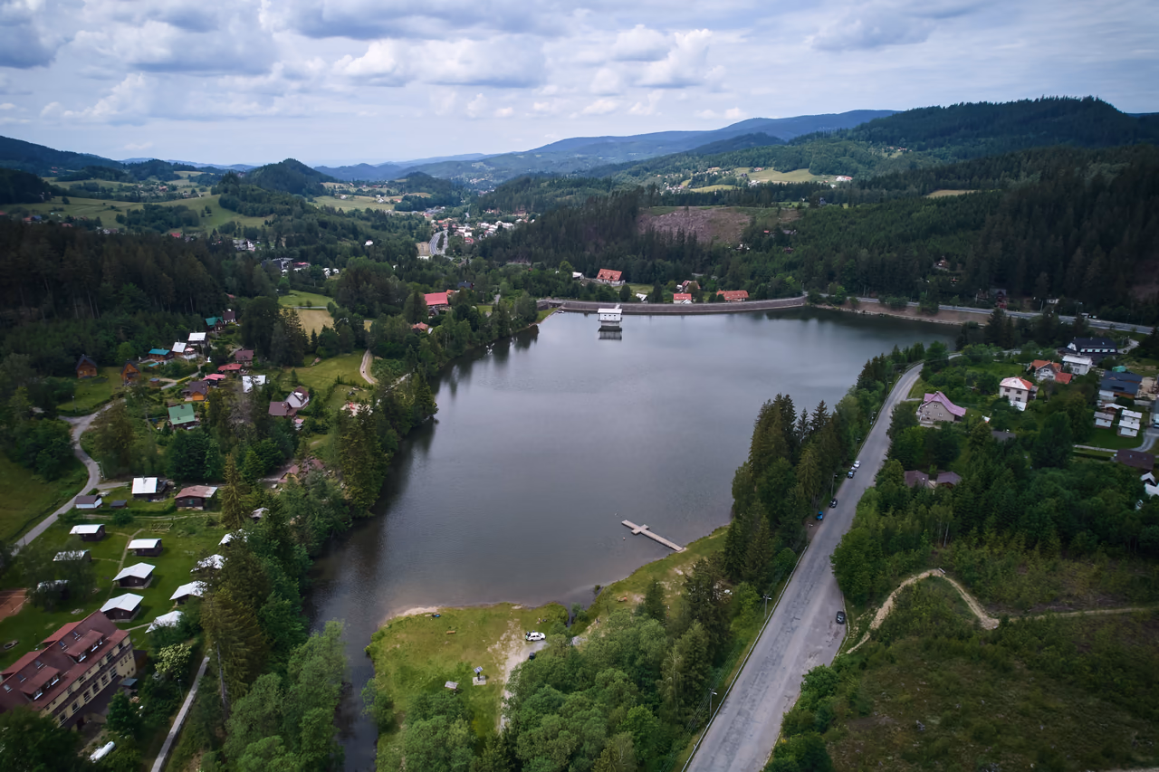 Blick über den Stausee zum Damm mit dem Turmbauwerk, im Vordergrund die Hütten des Camps zwischen den Bäumen und der Badesteg an der Liegewiese