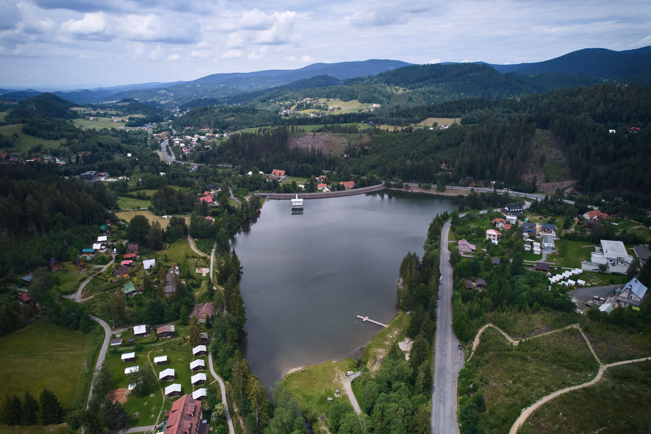 Der ganze Stausee Horní Bečva von oben - der Damm mit dem Turmbauwerk im Hintergrund, Badesteg und Liegewiese in der vorderen Bucht, die Straße am rechten Ufer entlang