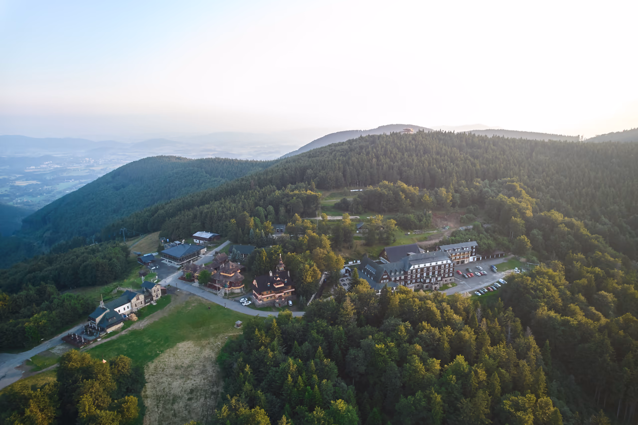 Letecký pohľad na sedlo Pustevien: drevené stavby pozdĺž hlavnej cesty, horský hotel s parkoviskom a trávnatá zjazdovka, v pozadí zalesnené hrebene a kraj v doline