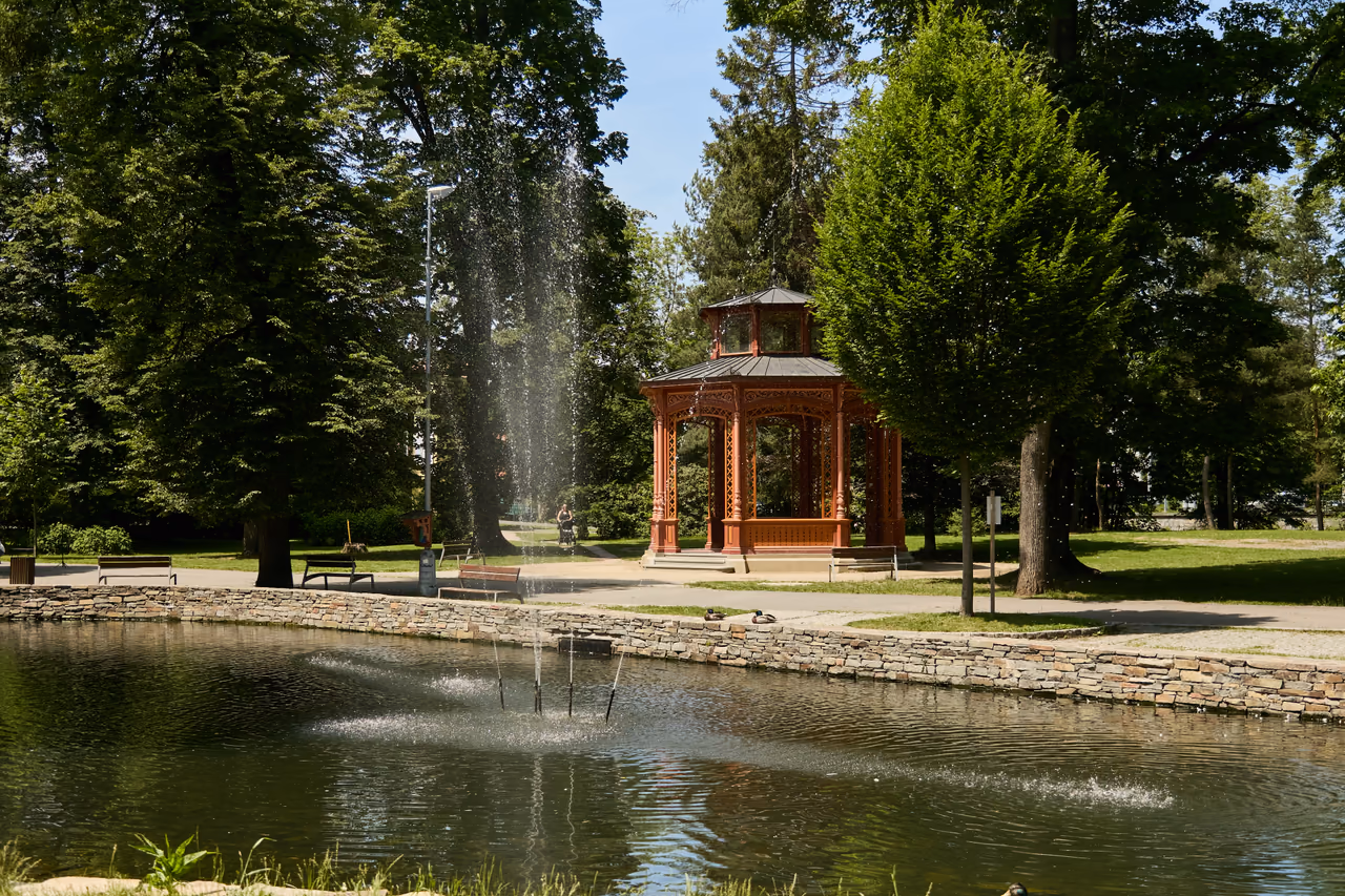 Jeziorko z fontanną w rožnovskim parku miejskim, w tle między drzewami altana muzyczna