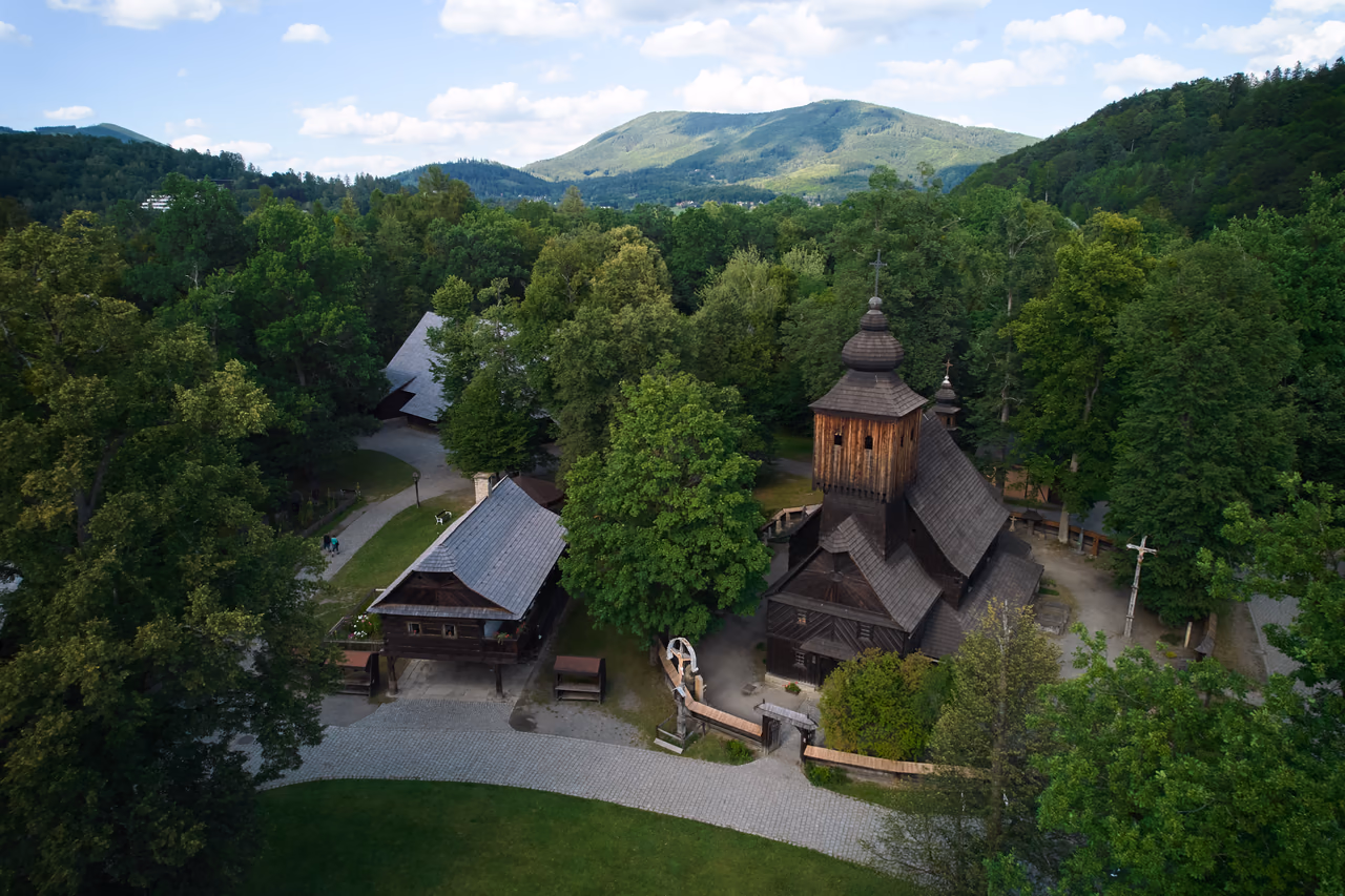 Luftbild der Kirche der hl. Anna zwischen den Bäumen des Dřevěné městečko, dahinter ein Beskidenkamm