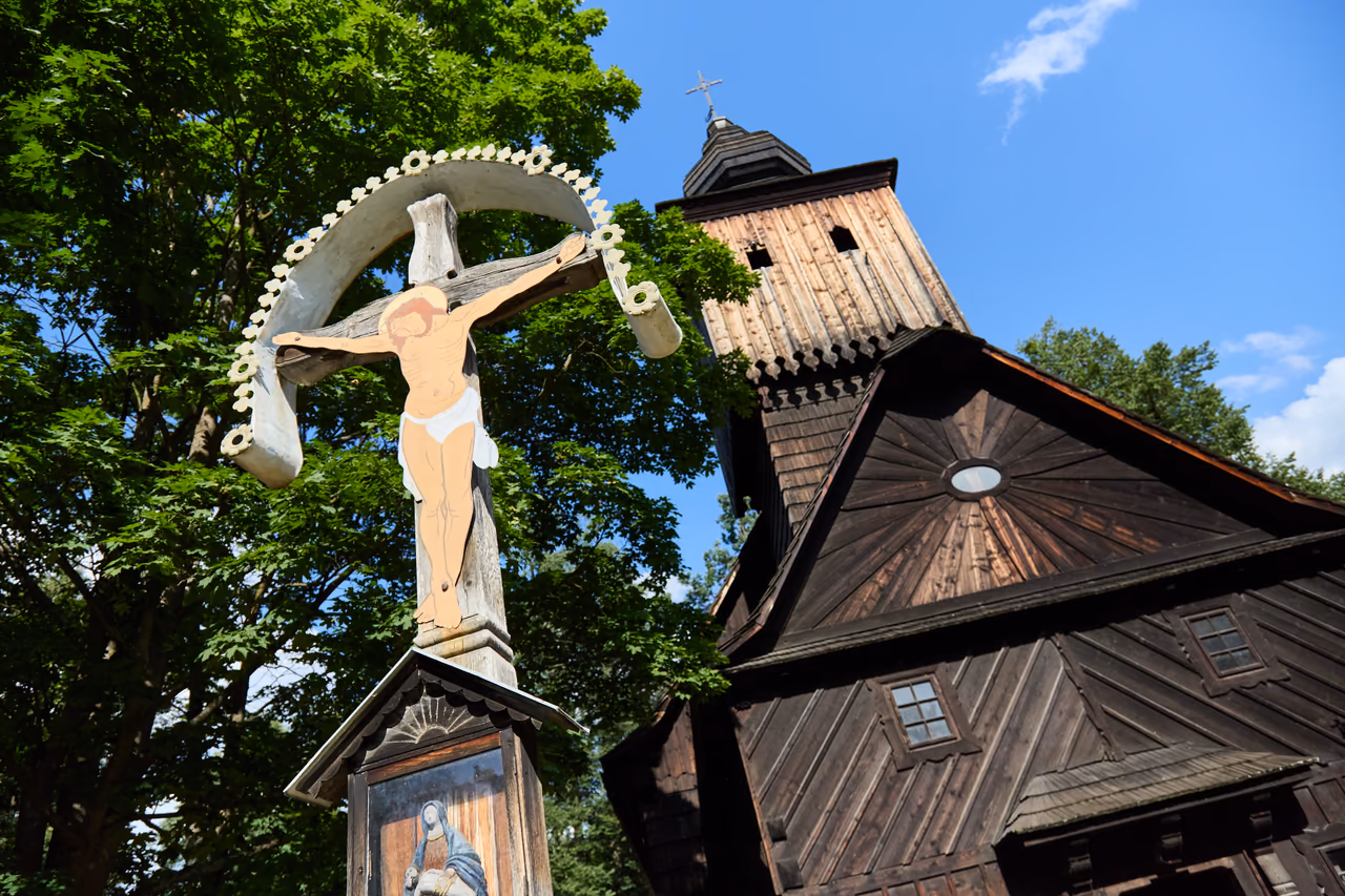 Kreuz mit dem Gekreuzigten unter einem geschmiedeten Bogen, dahinter der Schindelturm der Kirche der hl. Anna