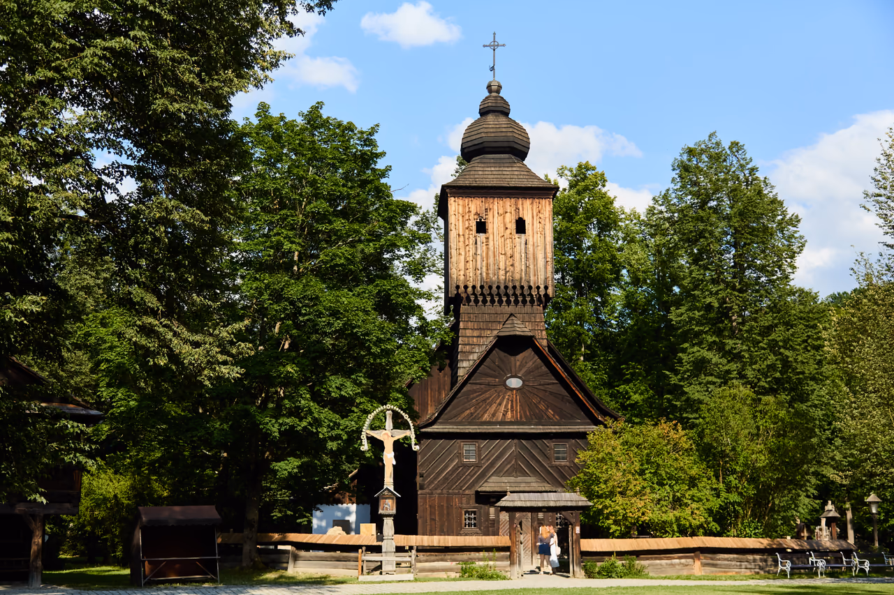 Fassade der Kirche der hl. Anna mit mächtigem Schindelturm, davor ein Kreuz mit dem Gekreuzigten
