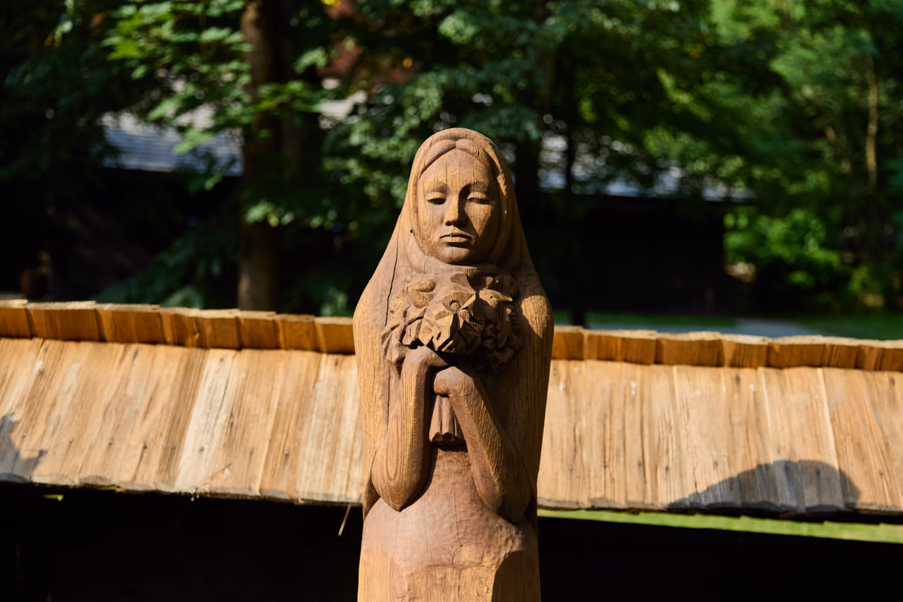 Geschnitzte Holzfigur einer Frau mit einem Blumenstrauß in den Händen, dahinter das Schindeldach der Umfassungsmauer