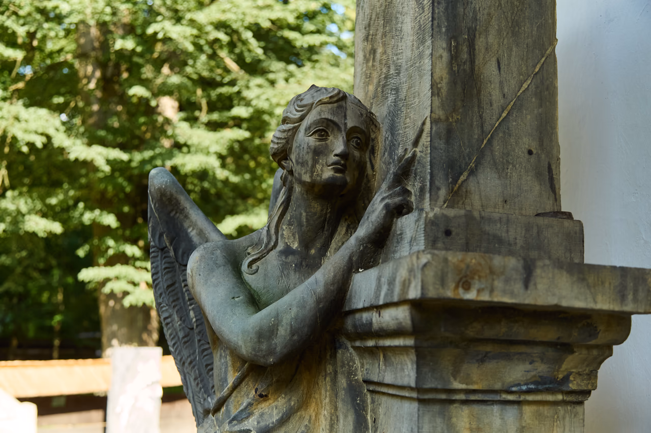 Metallfigur eines Engels mit grüner Patina, an eine Steinsäule gelehnt, die Hand weist nach oben